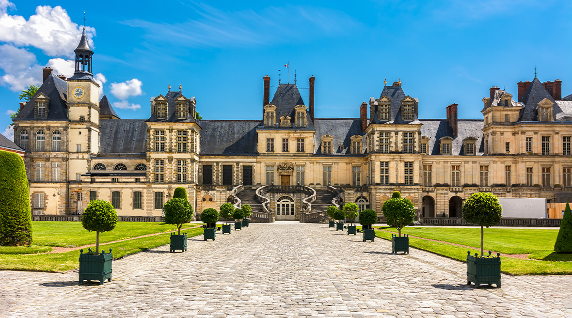 A large chateau with elegant gardens and a paved area on a summer’s day